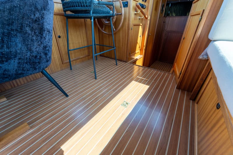De foto toont een interieur van een boot met een houten vloer en wanden, een stuurwiel, een barkruk en een bank.