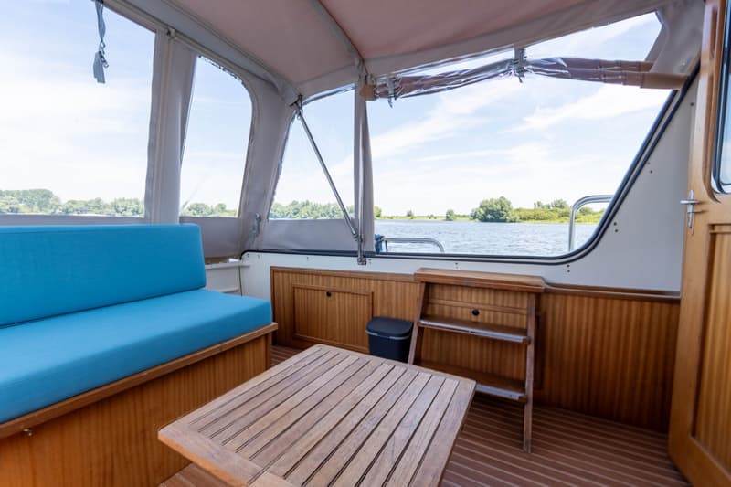 De foto toont het interieur van een boot met een blauwe bank, een houten tafel en uitzicht op water en groen.