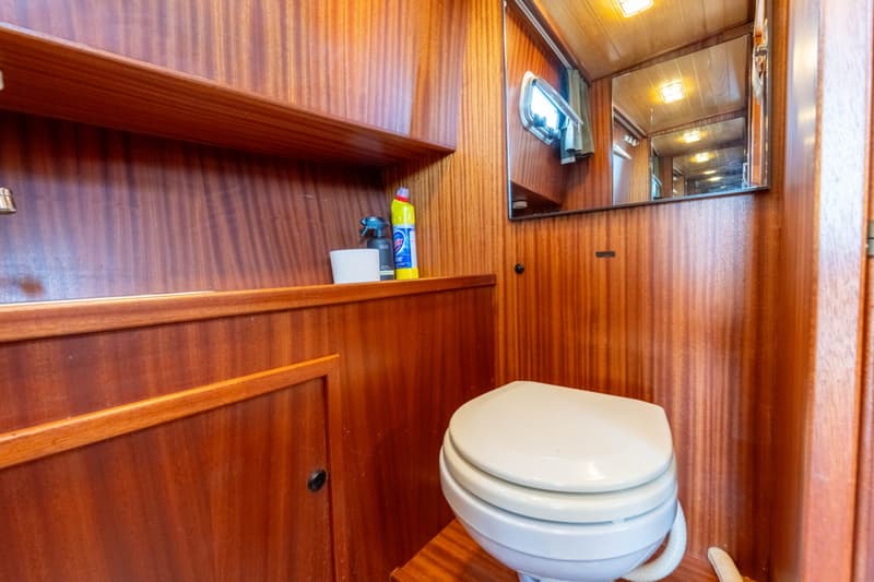 Een houten toiletruimte met een wit toilet, een spiegel, een patrijspoort en schoonmaakmiddelen op een plank.