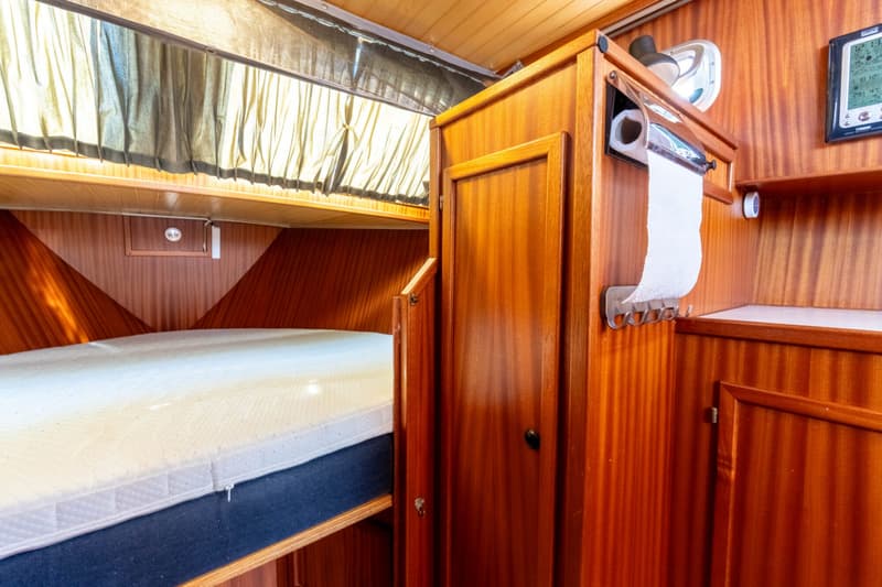 De foto toont een interieur van een boot met een bed, houten kasten, een papieren handdoekhouder en een weerstation.