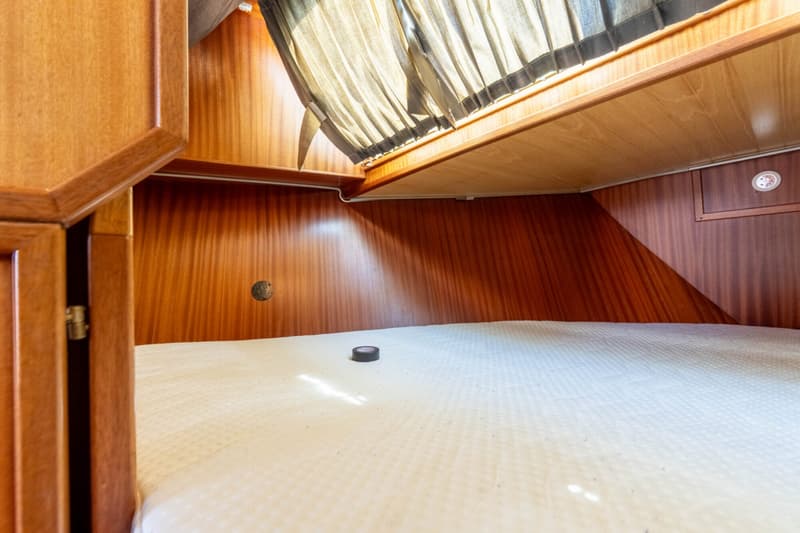 Een kajuit met houten wanden en kasten, een wit matras, een raam met gordijn en een ronde lamp.