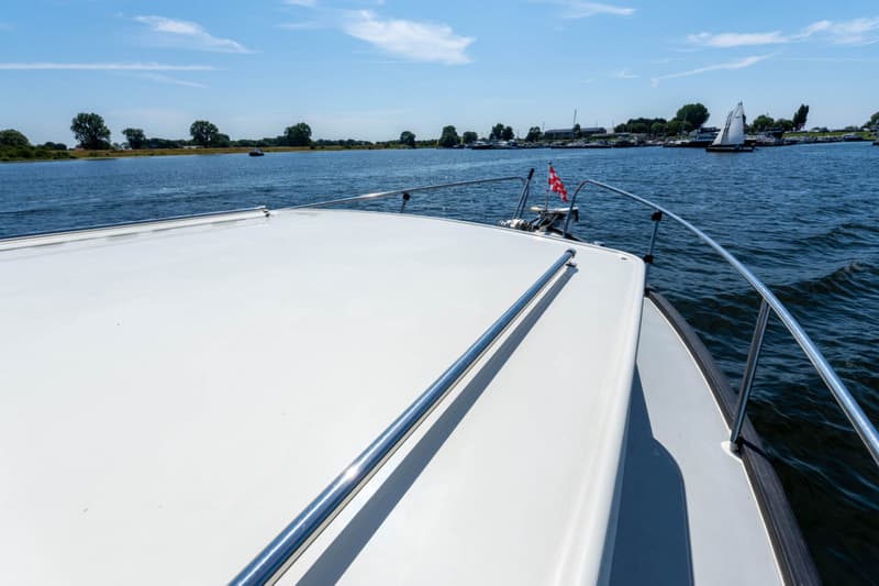 De foto toont de boeg van een witte boot met een rood-wit geblokte vlag, varend op water met andere boten en land in de…