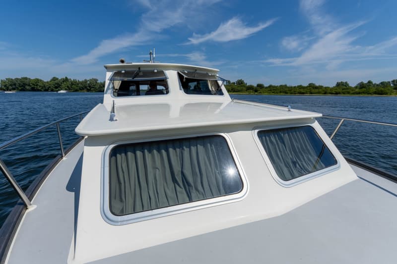 De foto toont de boeg van een witte motorboot met ramen en gordijnen, varend op een waterweg met bomen op de achtergrond.