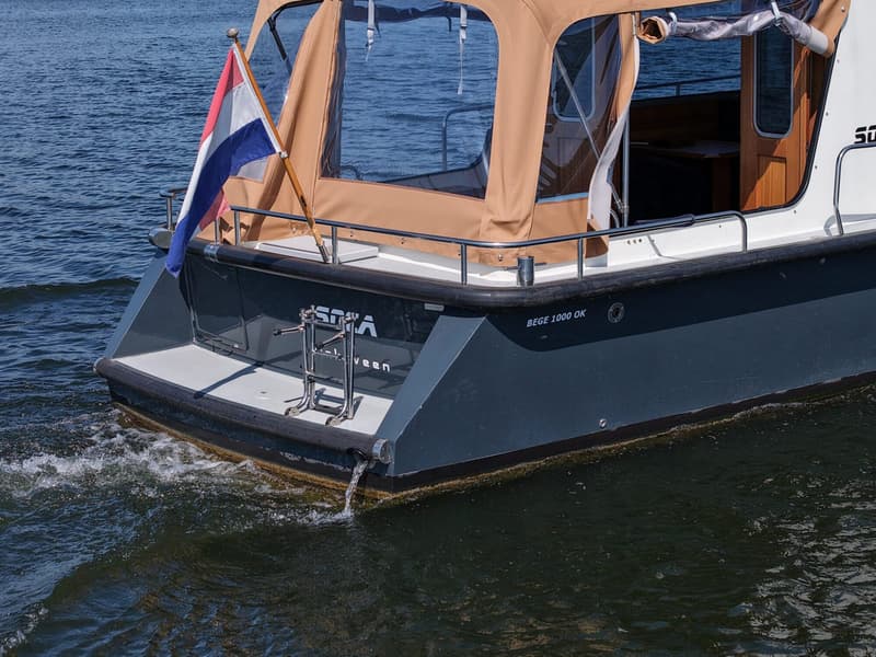 Een donkergrijze boot met een oranje kap, een Nederlandse vlag en de tekst "BEGE 1000 OK" op de zijkant.