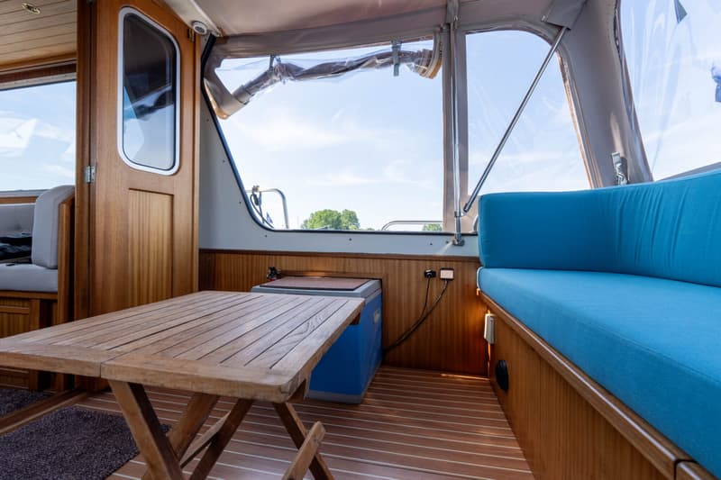 Een houten tafel en blauwe kussens op een boot met houten wanden en vloer, met uitzicht op water en bomen.
