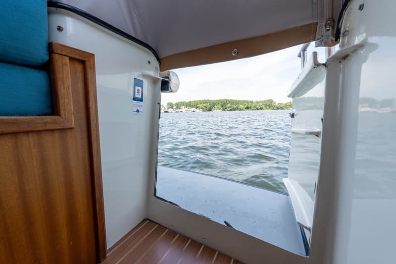 Een interieur van een boot met een houten deur, blauwe kussens en uitzicht op water met bomen en andere boten aan de horizon.