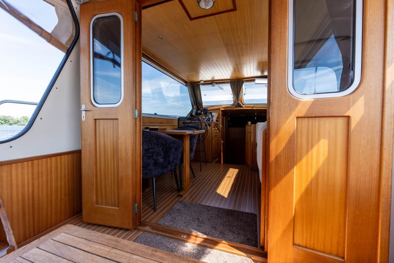 Een interieur van een boot met houten wanden en plafond, een stuurwiel en een tafel met stoelen.