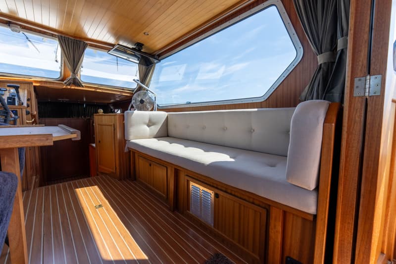 De foto toont het interieur van een boot met houten lambrisering, een lange bank met lichte kussens en grote ramen.