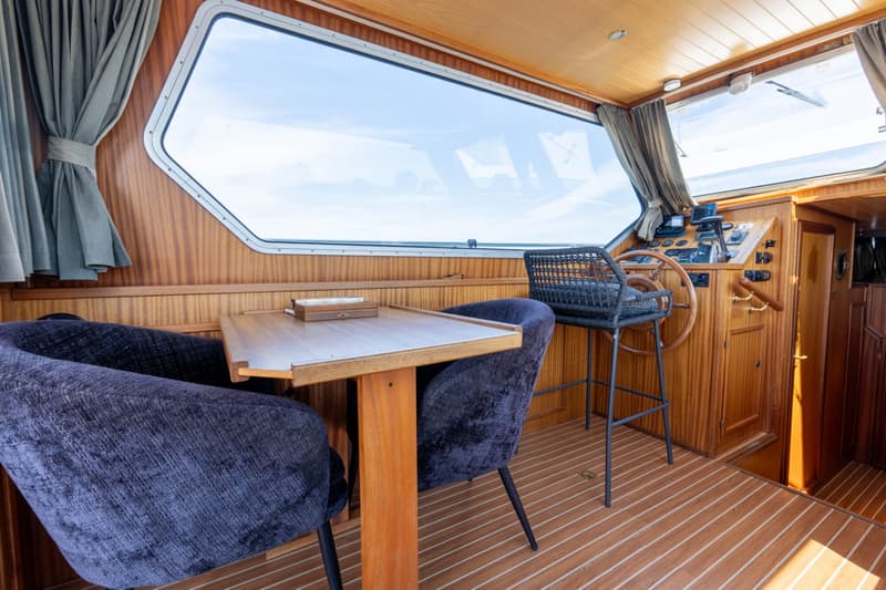Een interieur van een boot met houten wanden en vloer, een tafel met twee stoelen, en een stuurstand.