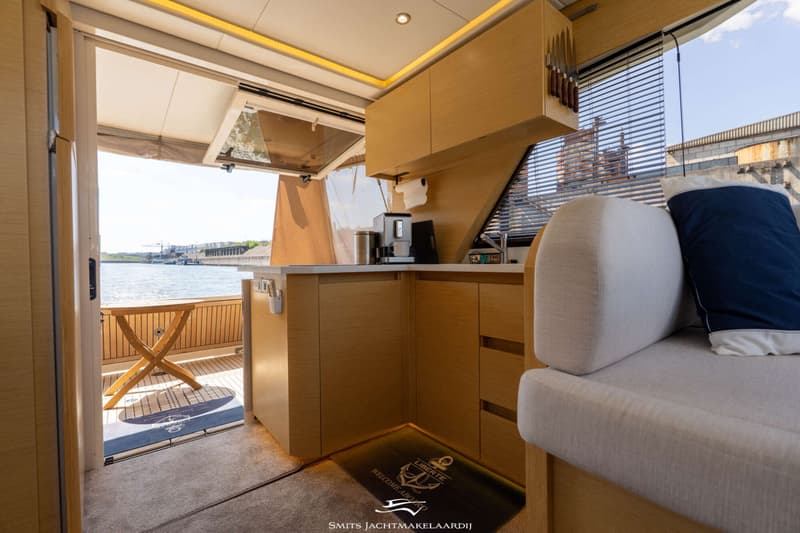 Een interieur van een boot met een keukenblok, een bank en uitzicht op water met een kade en gebouwen.