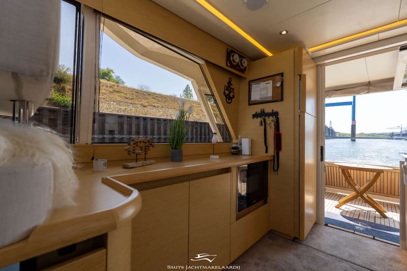 Een interieur van een boot met houten kasten, een raam met uitzicht op een dijk en een open deur naar het dek.
