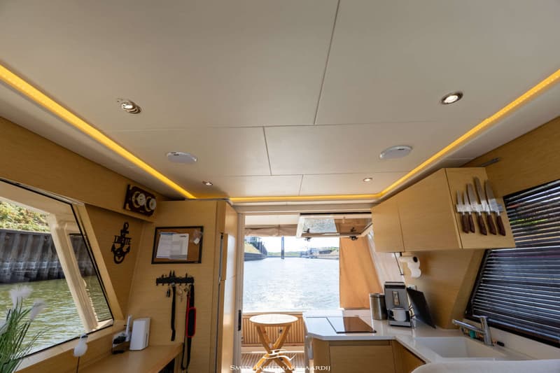 Een interieur van een boot met een keukenblok, houten kasten, een raam met uitzicht op water en een plafond met verlichting.