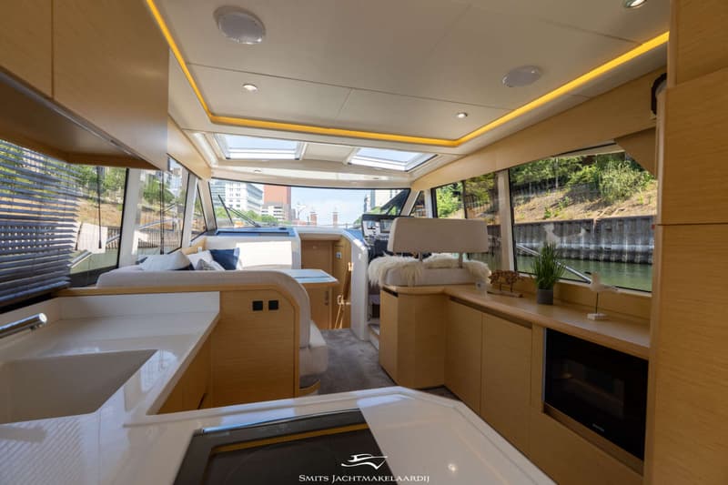 Een interieur van een boot met een keukenblok, zitgedeelte, stuurstand en ramen met uitzicht op water en bebouwing.