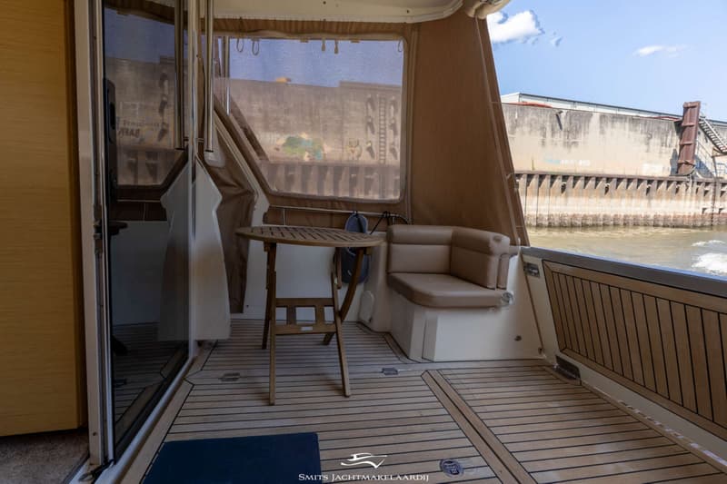 Een bootinterieur met een houten vloer, een beige bank, een houten tafel en een uitzicht op een waterweg met een betonnen…