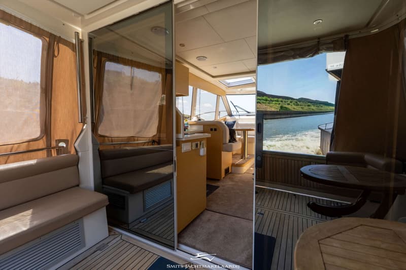 Een interieur van een boot met een glazen schuifdeur, banken, een tafel en uitzicht op water en landschap.