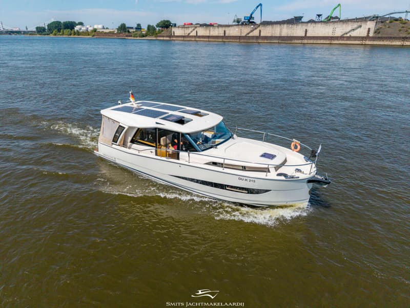 Een witte motorboot met zonnepanelen op het dak vaart over een rivier met industriële gebouwen op de achtergrond.