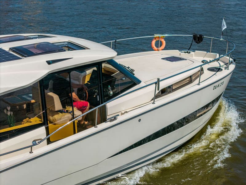 Een witte motorboot met een persoon aan het roer vaart over water, met zonnepanelen op het dak en een reddingsboei aan de…