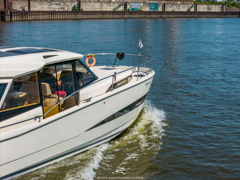 Een witte motorboot met een oranje reddingsboei en een vlag vaart op het water, met een kade op de achtergrond.