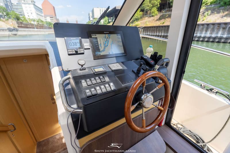 De foto toont een stuurstand met een houten stuurwiel, diverse knoppen, meters en een Raymarine navigatiescherm.