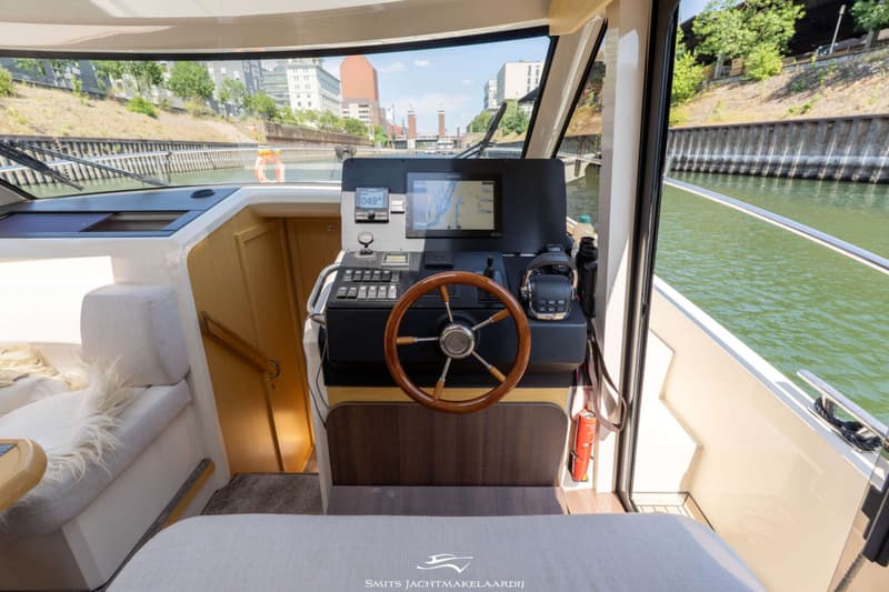 De foto toont het stuurhuis van een boot met een houten stuurwiel, navigatiescherm en bedieningspanelen.