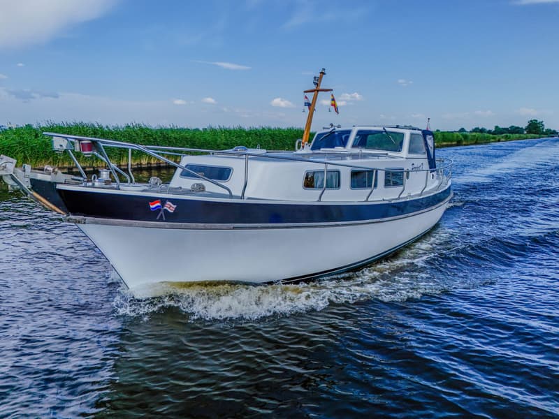 Een witte motorboot met een donkerblauwe romp vaart op een waterweg met rietkragen onder een blauwe lucht.