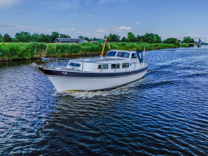 Een witte motorboot met een donkere romp vaart over een waterweg met groene oevers en een blauwe lucht.