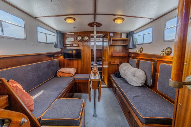 De foto toont de kajuit van een boot met houten wanden, grijze banken met oranje biezen, en een inklapbare tafel.