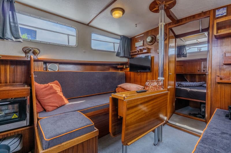 De foto toont het interieur van een boot met houten wanden, een bank met grijze kussens en oranje biezen, en een opklapbare…