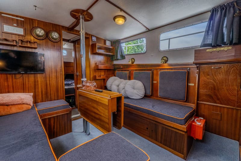 De foto toont een houten interieur van een boot met zitbanken, een tafel, een televisie en ramen.