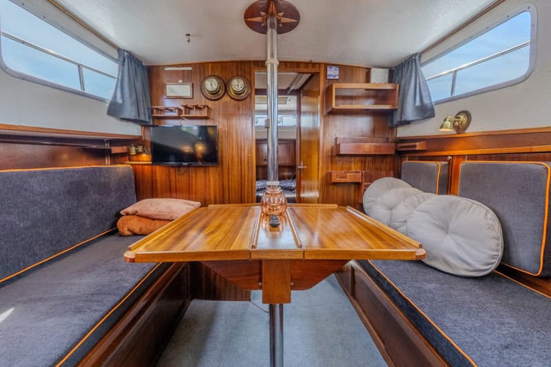 De foto toont het interieur van een boot met een houten tafel, banken met grijze kussens en ramen aan beide zijden.