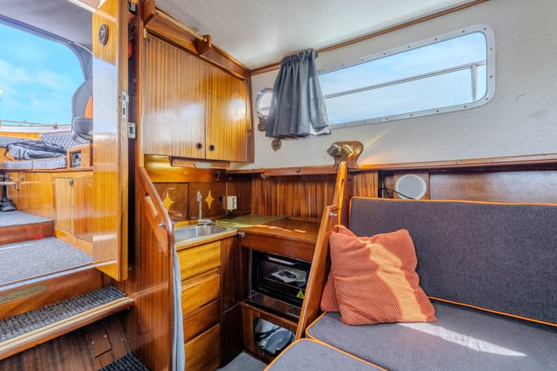 De foto toont het interieur van een boot met houten afwerking, een kombuis, een zithoek en een trap naar een hoger dek.