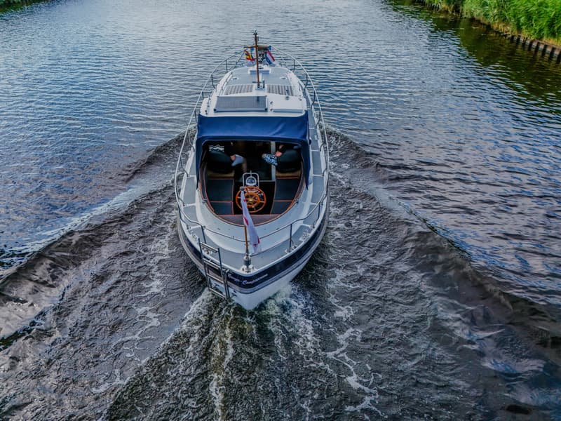 Een boot met een blauwe kap en een houten stuurwiel vaart over water, gezien van bovenaf.