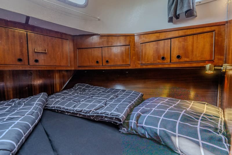 Een kajuit met een bed met donkergrijze dekens en kussens met een wit rasterpatroon, omringd door houten kasten.