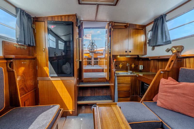 De foto toont het interieur van een boot met houten afwerking, een kombuis, zitbanken en een doorgang naar de stuurstand.