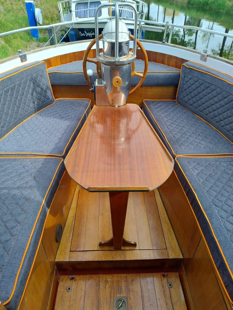 Een houten bootinterieur met een tafel, stuurwiel, kompas en grijze kussens met oranje biezen.