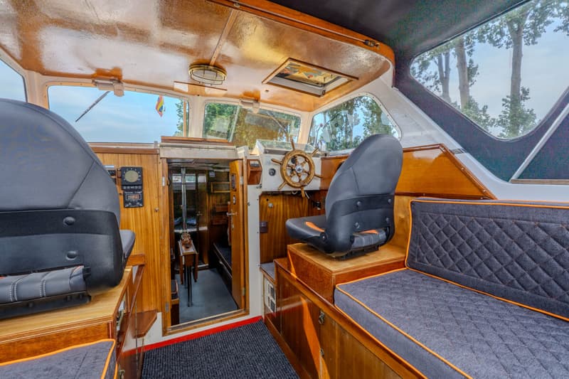 De foto toont het interieur van een boot met houten afwerking, een stuurwiel, twee stoelen en een bankje.
