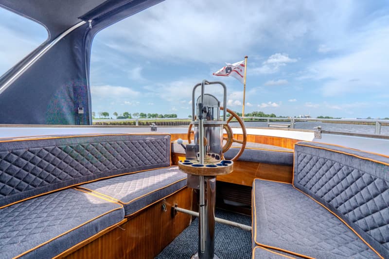 Een bootinterieur met grijze kussens met oranje biezen, een houten stuurwiel en een vlag met een ster en een 'B'.