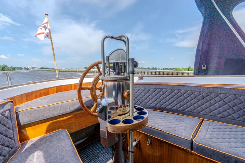 Een houten stuurwiel en een kompasstandaard met bekerhouders zijn zichtbaar op een boot met grijze kussens en een vlag.