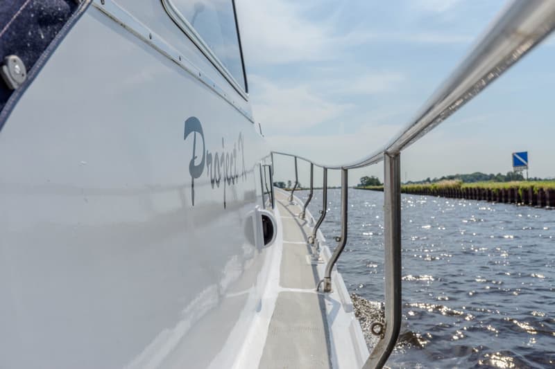 Een witte boot met de tekst "Project 2" op de zijkant vaart over water met een reling aan de rechterkant.