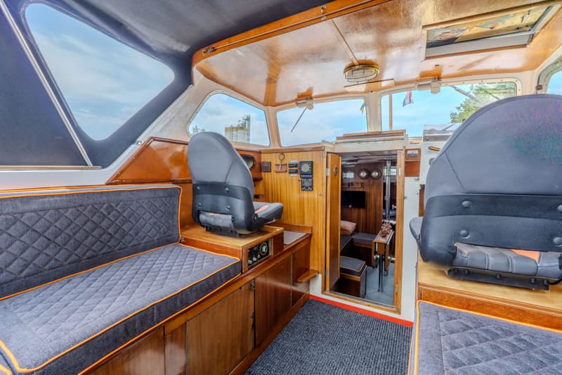 De foto toont het interieur van een boot met houten afwerking, twee grijze stoelen en een bank met oranje biezen.