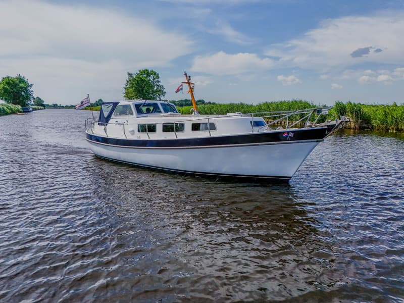 Een witte motorboot met een donkere stootrand vaart op een kanaal met rietkragen en bomen langs de oevers.
