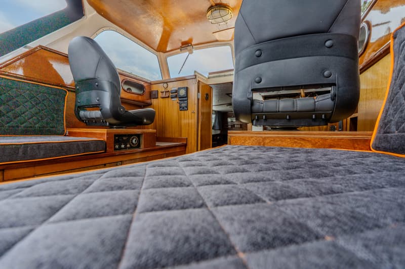 De foto toont het interieur van een boot met houten wanden en plafond, grijze gestoffeerde banken en twee zwarte stoelen.
