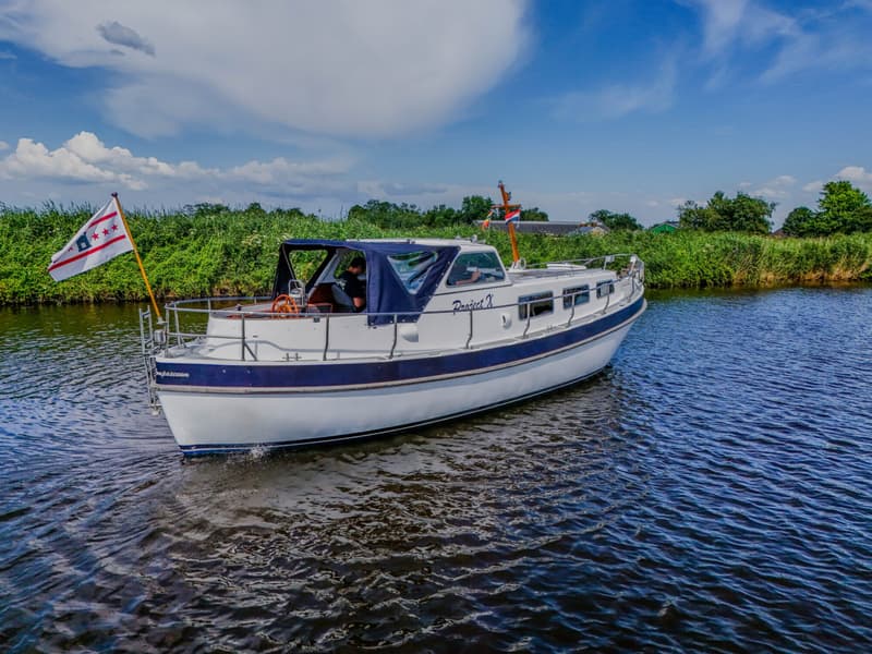 Een witte boot met blauwe accenten en de naam "Project X" vaart op een waterweg met groene oevers onder een blauwe lucht.