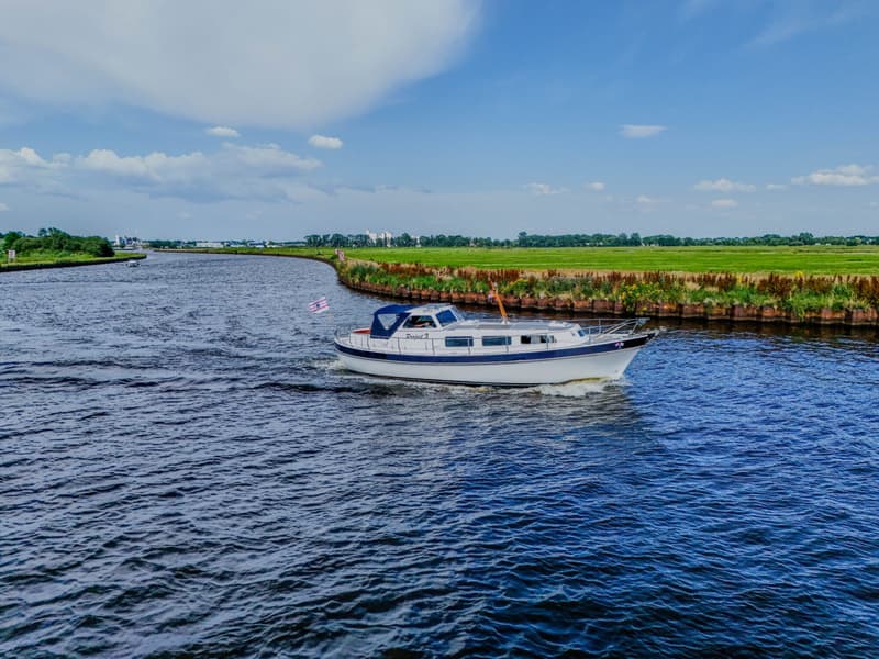 Een witte motorboot met blauwe accenten vaart op een rivier met groene oevers onder een blauwe lucht met wolken.
