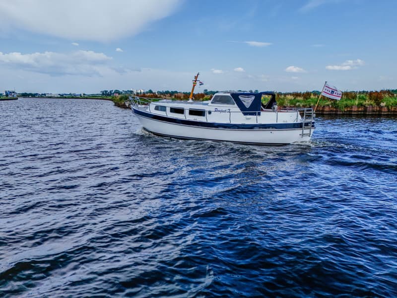Een witte motorboot met blauwe accenten en de naam "Project X" vaart op een waterweg onder een bewolkte hemel.