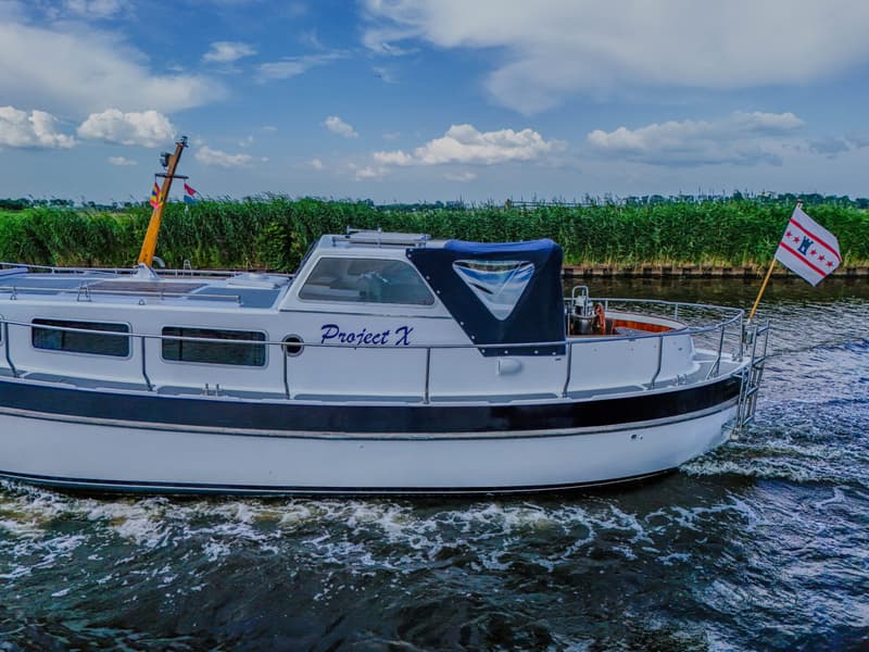 Een witte motorboot met de naam "Project X" vaart op een kanaal langs een rietkraag onder een bewolkte hemel.