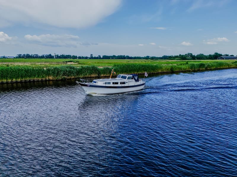 Een witte motorboot met blauwe accenten vaart op een kanaal met groene oevers onder een blauwe lucht met wolken.