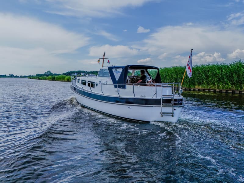 Een witte motorboot met blauwe accenten vaart op een waterweg met rietkragen onder een bewolkte lucht.