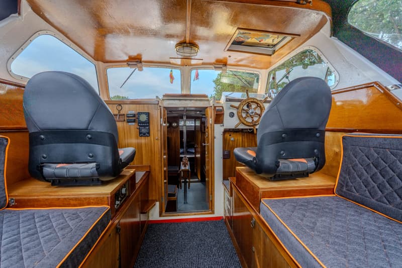 De foto toont het interieur van een boot met houten afwerking, twee stuurstoelen, een stuurwiel en een doorgang naar een…