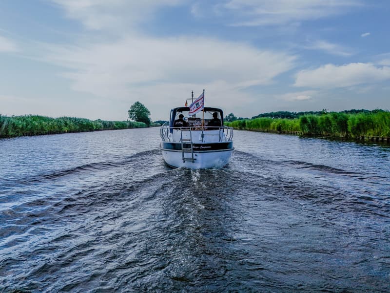 Een witte motorboot met een donkerblauwe romp vaart over een waterweg, omringd door groene oevers onder een bewolkte lucht.